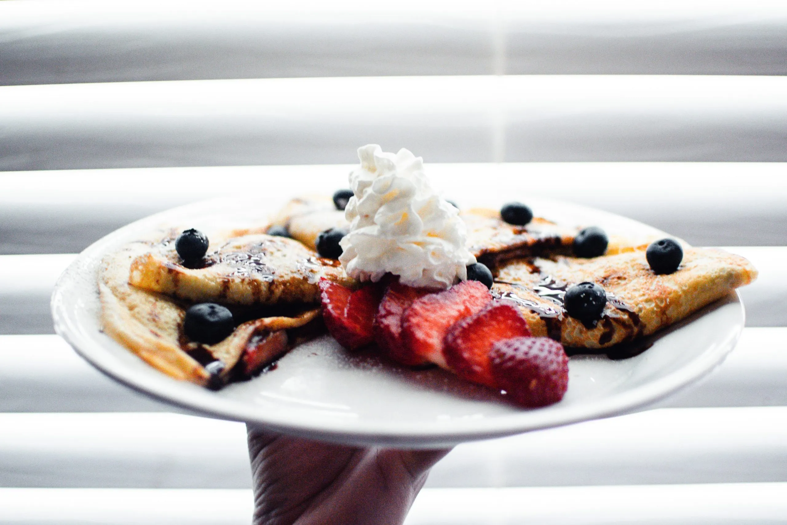 A hand holding a plate of crepes topped with whipped cream, blueberries and strawberries