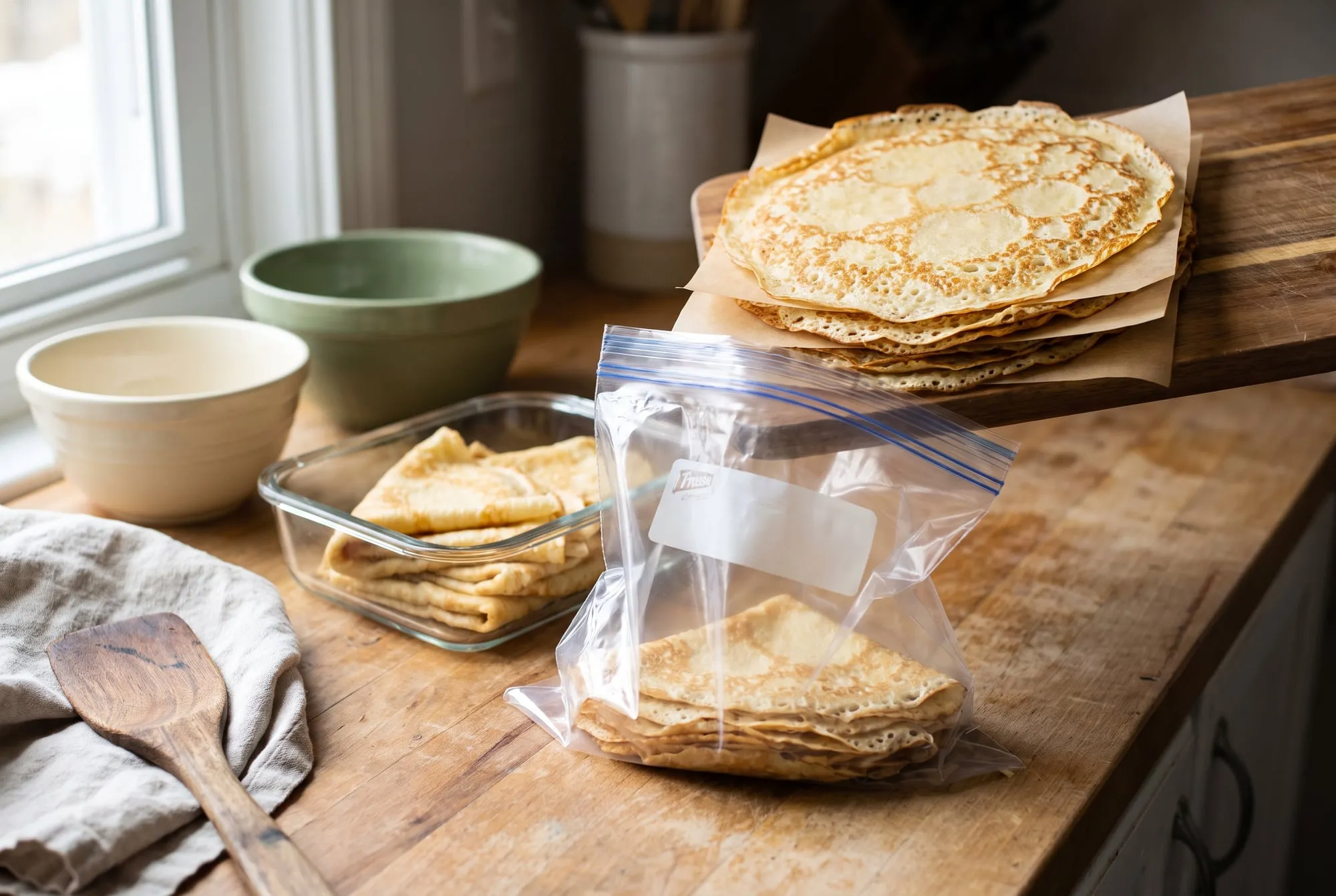 A stack of crepes layered with parchment paper going into a freezer bag, with a container of folded crepes beside it